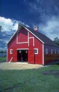 A photo of a red barn with white trim, barn doors open; sitting against a blue sky with fluffy white clouds.